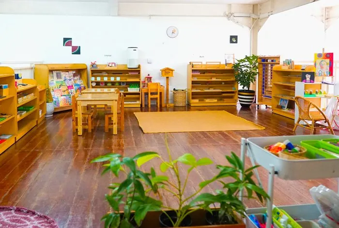 A prepared Montessori classroom with low open shelves, tables and a work rug